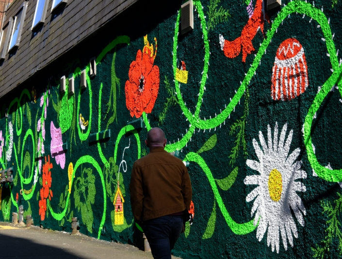 Image shows the Reid Lane mural with a community garden theme. The artwork contains local community garden flowers and many animals including snails, birds, ladybirds and cats. Mural by Ida Henrich